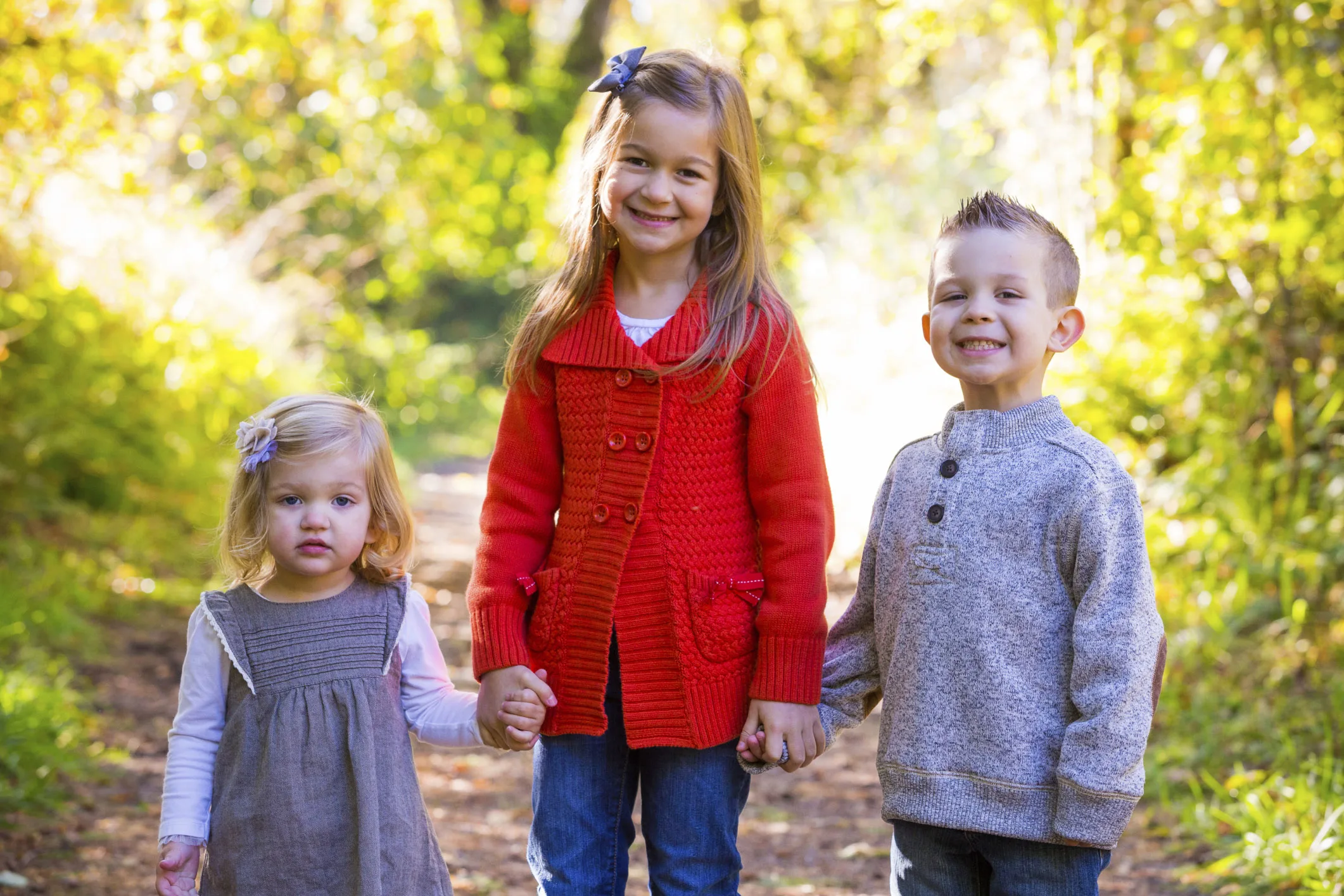 Three smiling children standing on a sunlit path in a park, wearing fall clothing.