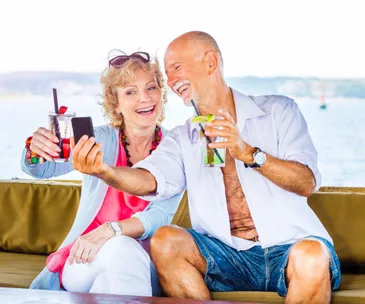 Elderly couple on a boat smiling, taking a selfie, enjoying drinks with a scenic ocean view in the background.