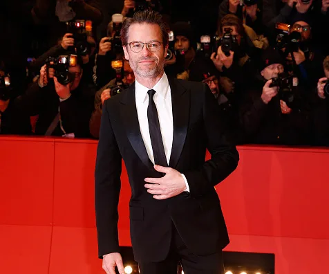 Guy Pearce in a formal suit at a red carpet event, photographers in the background capturing the moment.