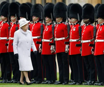 A woman in a white coat inspects a line of guards in red uniforms and bearskin hats.