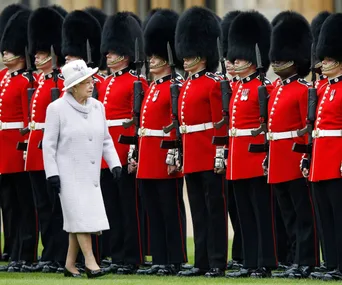 A woman in a white coat inspects a line of guards in red uniforms and bearskin hats.