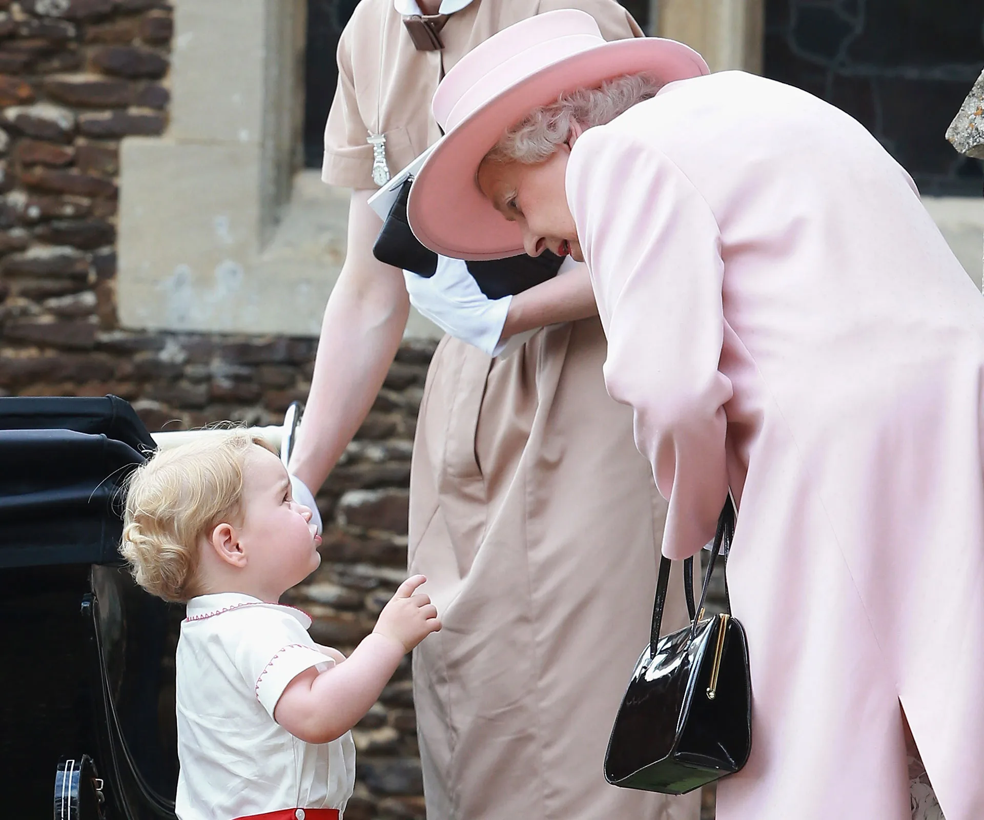 A young boy in a white outfit looks up at an elderly woman in a pink coat and hat, sharing a tender moment outside.