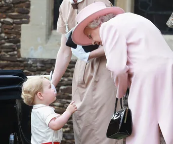 A young boy in a white outfit looks up at an elderly woman in a pink coat and hat, sharing a tender moment outside.
