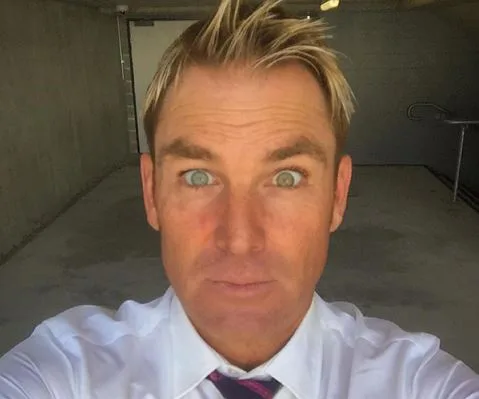 A man with tousled blonde hair and a surprised expression takes a selfie in a concrete room.