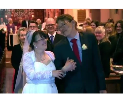 Bride in a white dress and groom in a suit smiling and walking down the aisle amid applauding guests in a church.
