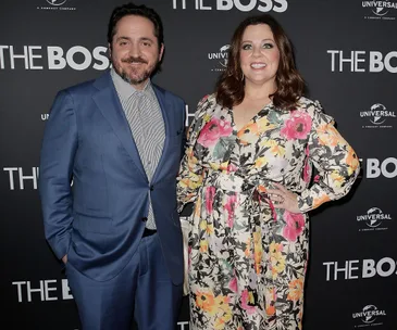 A couple posing at "The Boss" movie premiere, wearing a blue suit and a floral dress, smiling in front of a branded backdrop.
