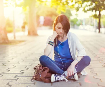 Teen girl sitting on a sidewalk wearing headphones, looking thoughtful and holding a phone.
