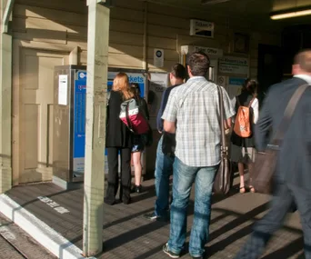 People standing in line at a train station ticket machine, with backpacks and bags.