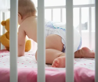 Baby in a diaper crawling in a crib with a white railing, with a teddy bear and polka dot bedding.