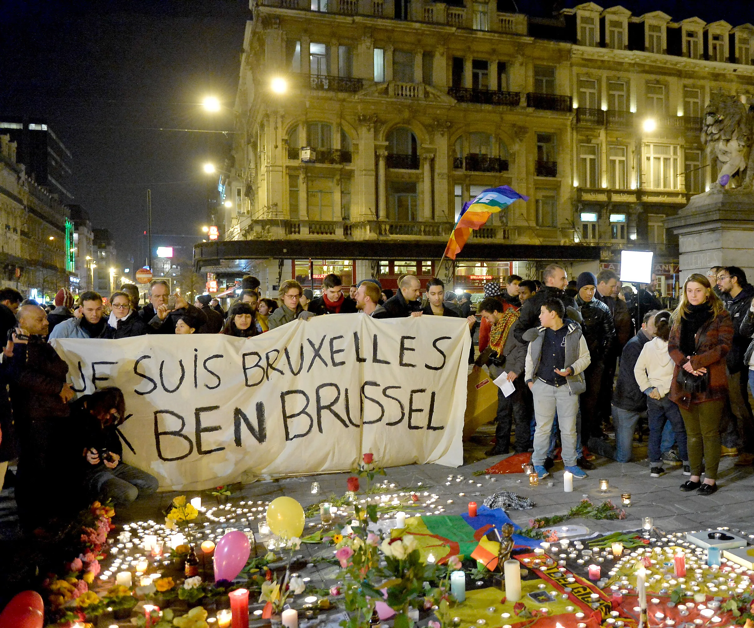 People gather at night in Brussels, holding a banner reading "JE SUIS BRUXELLES." Candles and flowers on the ground.
