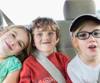 Three children smiling and posing in a car seat, one wearing glasses and a cap, enjoying a backseat travel adventure.