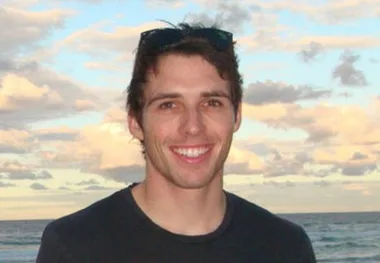 A person with short hair smiling broadly at the camera, ocean and cloudy sky in the background.