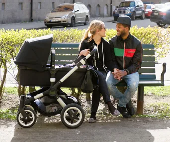 Couple sitting on a bench with a black pram beside them, enjoying a sunny day.
