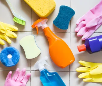 Cleaning supplies arranged on a tiled floor, including gloves, sponges, a brush, and spray bottles.