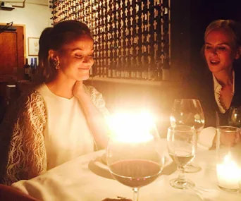 Reese Witherspoon smiling at birthday candles, sitting at a dimly-lit restaurant table with wine glasses.