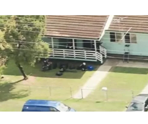 Aerial view of a vacant house with a front yard, several items on the porch, and vehicles parked nearby.