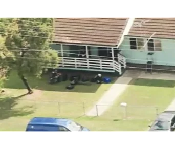 Aerial view of a vacant house with a front yard, several items on the porch, and vehicles parked nearby.