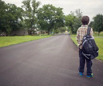 Young boy with a backpack stands alone on a rural road, facing away, surrounded by greenery and distant houses.