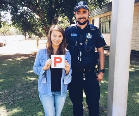 Young woman holds a 'P' plate next to a smiling police officer outside on a sunny day.