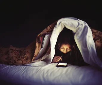 Child using smartphone under blanket at night, screen illuminating face.