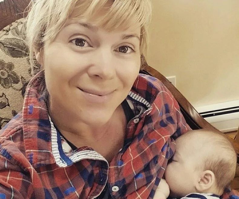 Woman in a plaid shirt breastfeeding a baby while smiling at the camera.