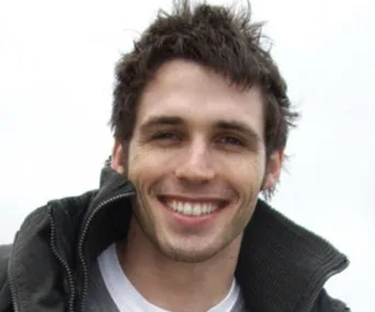 A smiling man wearing a dark jacket, with a blurred neutral background.