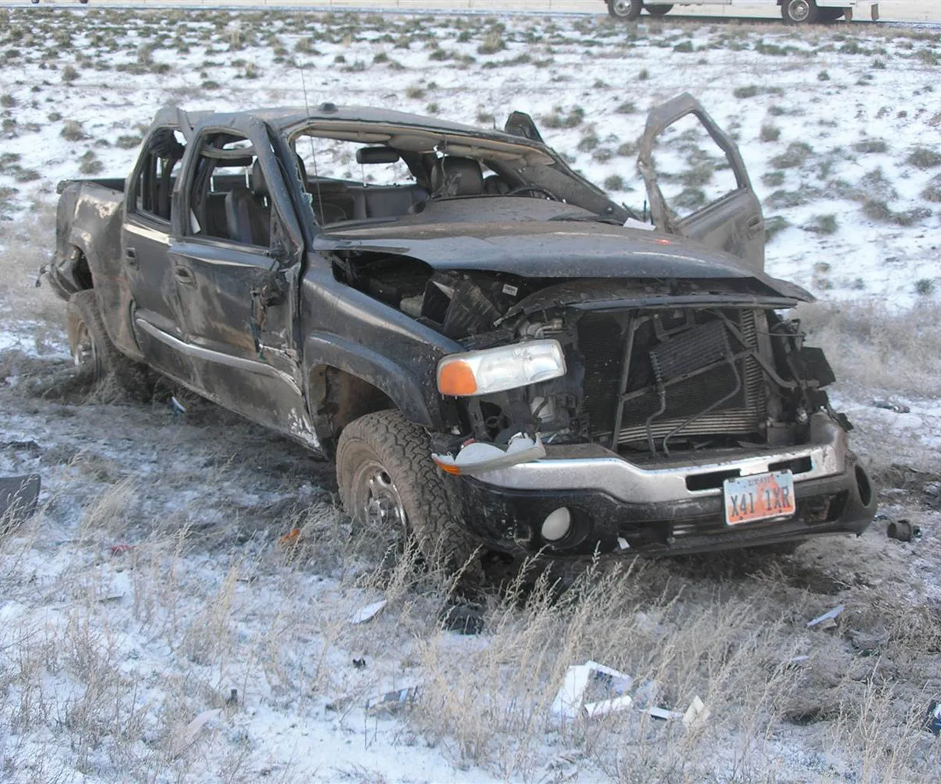 A severely damaged black pickup truck sits in a snowy field after a crash, with open doors and a crumpled front end.