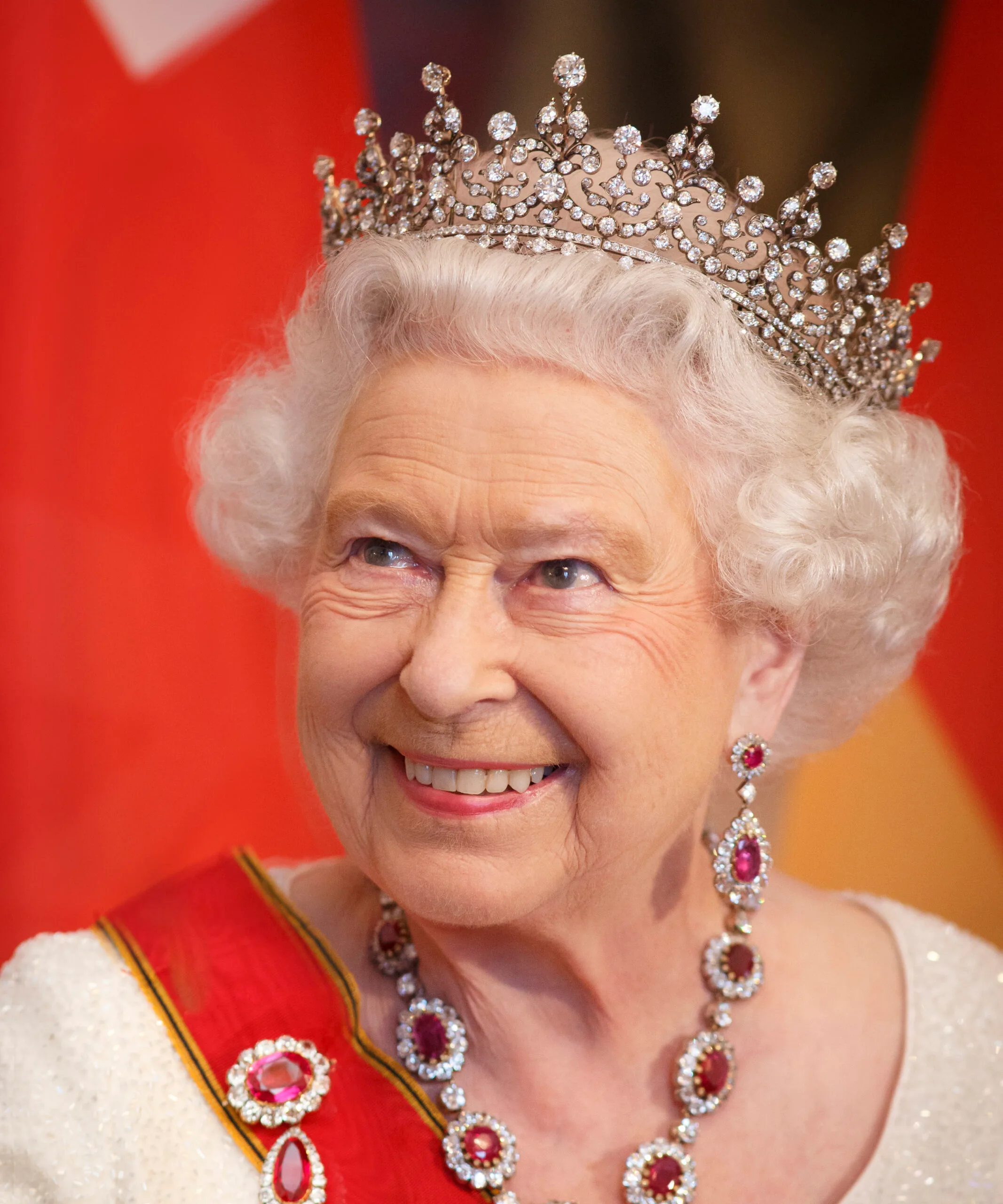 A smiling elderly woman wearing a jeweled tiara and red-and-white attire with matching necklace and earrings.