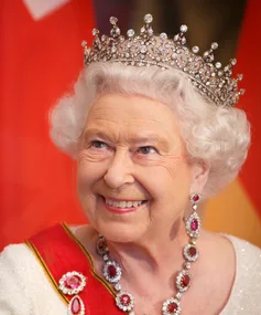 A smiling elderly woman wearing a jeweled tiara and red-and-white attire with matching necklace and earrings.