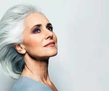 Elderly woman with gray hair and subtle makeup looking upwards against a light background.