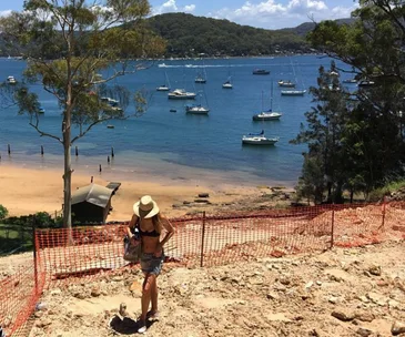 A woman in a hat and swimsuit stands on a rocky terrain by a beach, with sailboats and hills in the background.