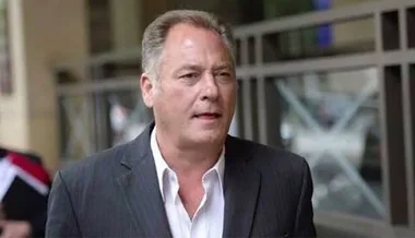 A man in a suit walks outside a building, appearing serious.