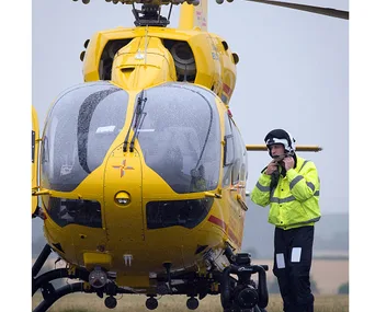 Helicopter pilot in high-visibility jacket stands beside a bright yellow rescue helicopter on a grassy field.