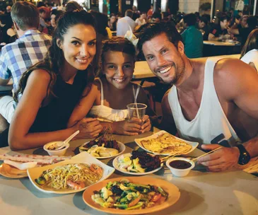 Family dining at a bustling Singaporean street food market, smiling with traditional dishes on the table.