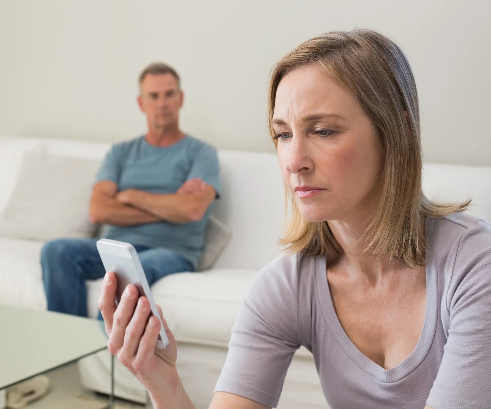 Woman focused on phone screen, man sits in background on couch with arms crossed, expressing concern or disconnect.