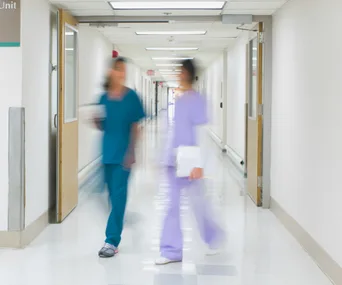 Blurry image of two nurses walking in a hospital corridor, one in blue scrubs and the other in purple, holding a clipboard.