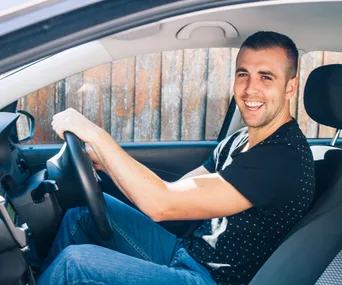 Man smiling while sitting in a car, wearing a black shirt and jeans, holding the steering wheel.