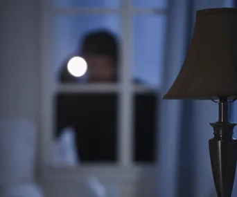 A blurred figure outside a window shines a flashlight into a dimly lit room, with a lamp visible in the foreground.