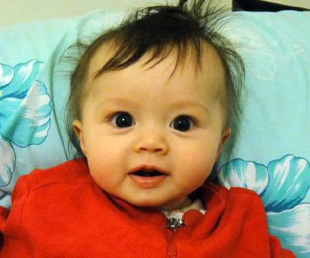 A baby with big eyes and dark hair, wearing a red outfit, is lying on a pillow with blue and white flowers.