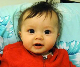 A baby with big eyes and dark hair, wearing a red outfit, is lying on a pillow with blue and white flowers.