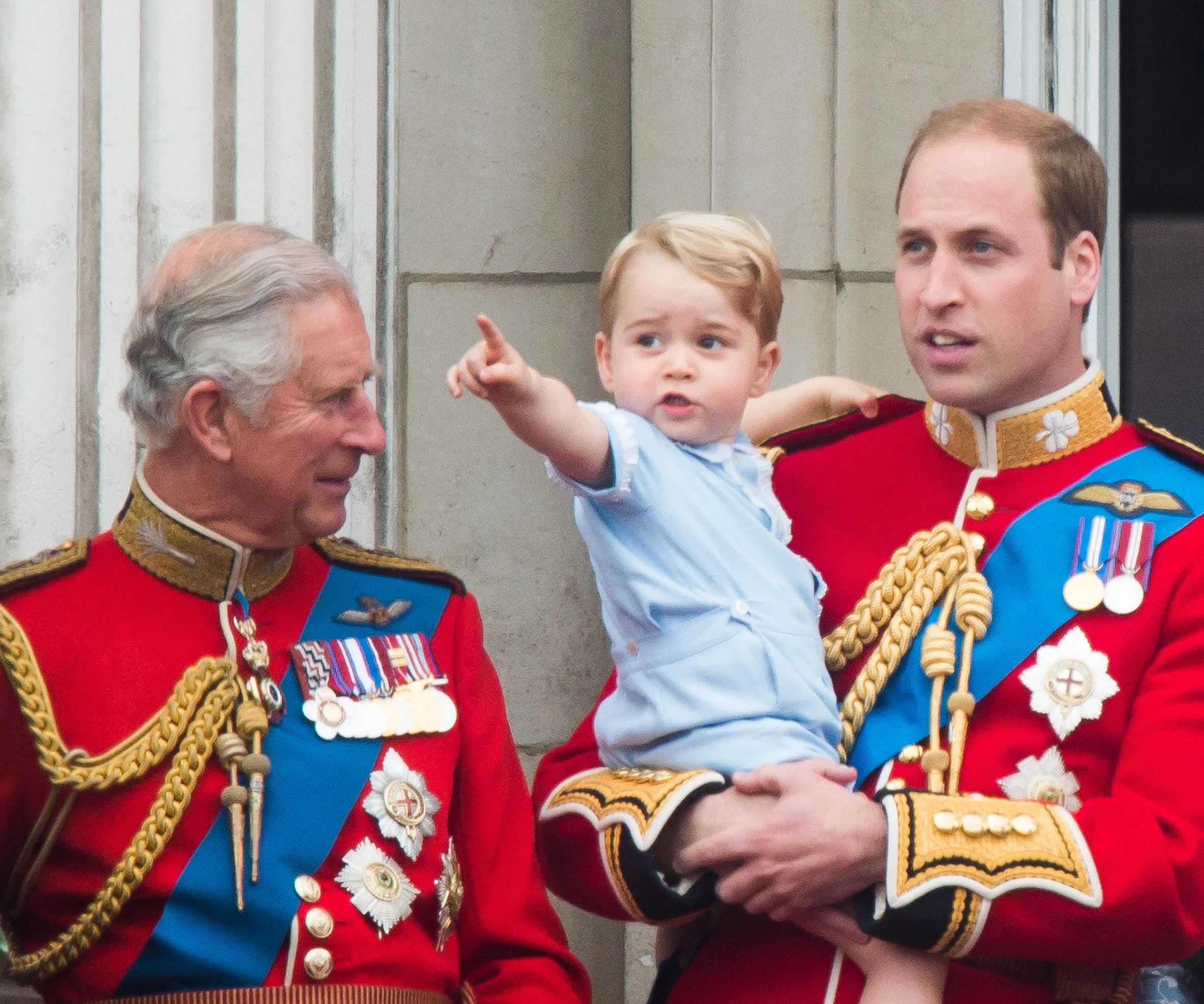 A child is pointing while being held by an adult in ceremonial military attire, with another adult looking on and smiling.
