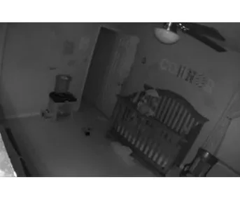 Nanny cam footage of a dimly lit nursery with a toddler standing eerily in a crib.