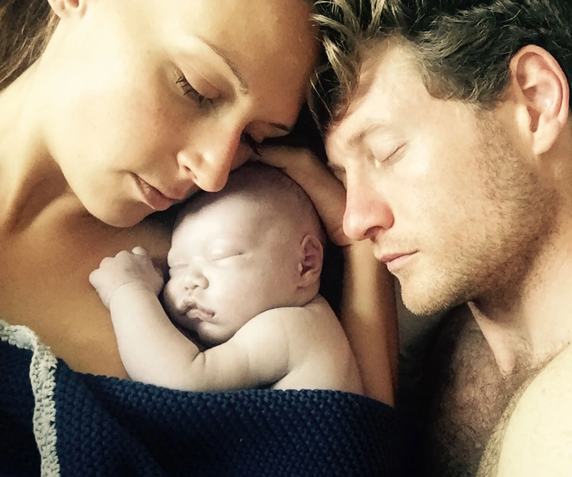Parents cuddling newborn baby wrapped in a blanket, all with peaceful expressions, highlighting tender family bond.