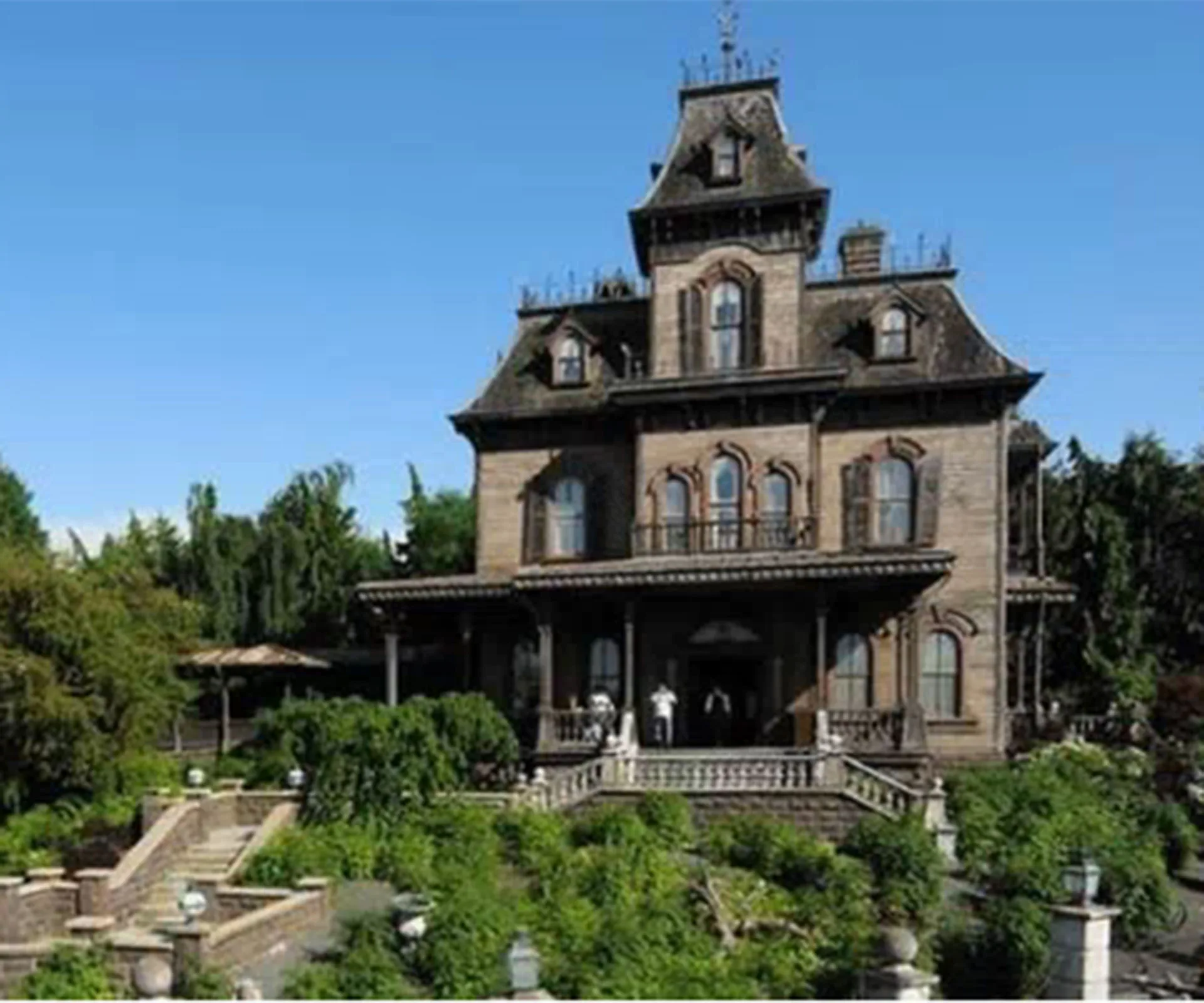 A large, old-fashioned mansion surrounded by greenery under a clear blue sky.