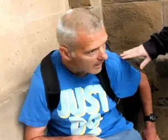 A man in a blue shirt with "JUST DO" is sitting against a wall, looking surprised as a hand reaches toward him.