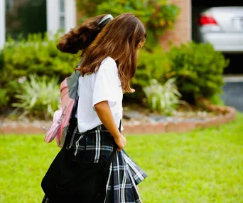 Two young girls in school uniforms walking on grass, wearing backpacks.