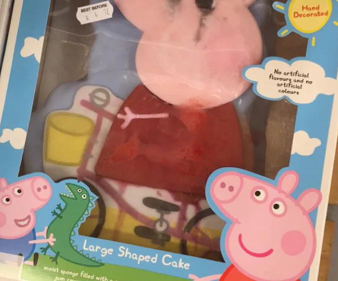 A Peppa Pig-themed cake with a misshaped face, in a colorful box featuring cartoon images of Peppa and a dinosaur.