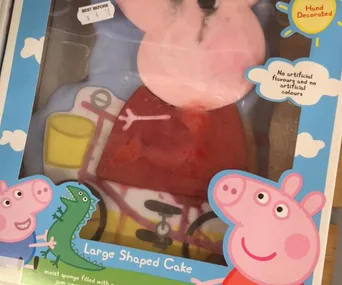 A Peppa Pig-themed cake with a misshaped face, in a colorful box featuring cartoon images of Peppa and a dinosaur.