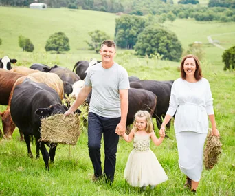 Man and woman holding hands with a child in a field, carrying hay, surrounded by cows.