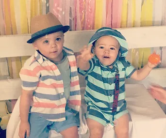 Two toddlers wearing striped outfits and hats sit on a bench with colorful streamers in the background.